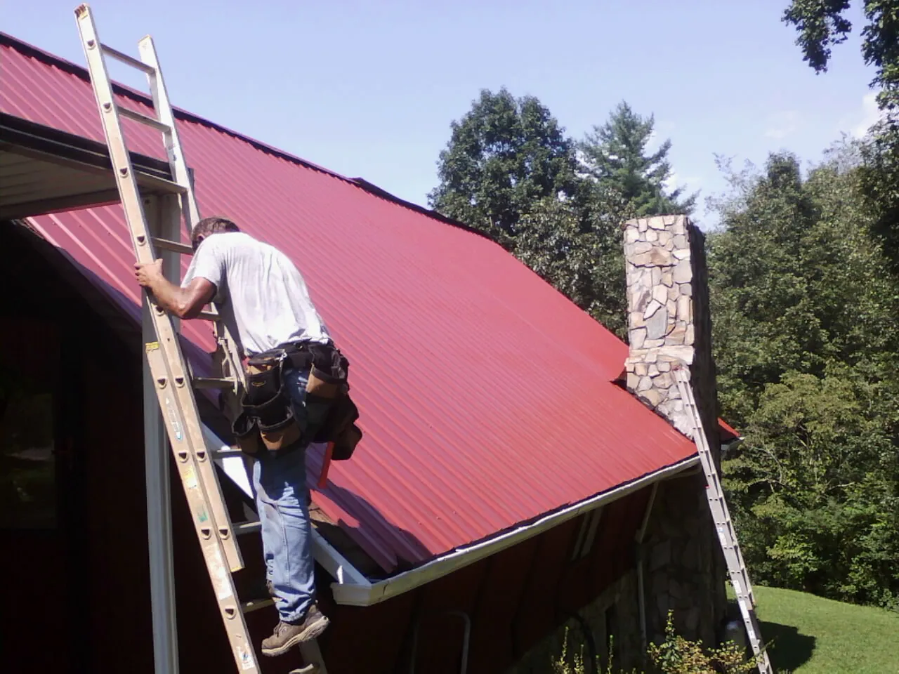 Roofer on a ladder beside a red metal roof and stone chimney — Soffit & Fascia Repair by Roofing Brothers in Muncie
